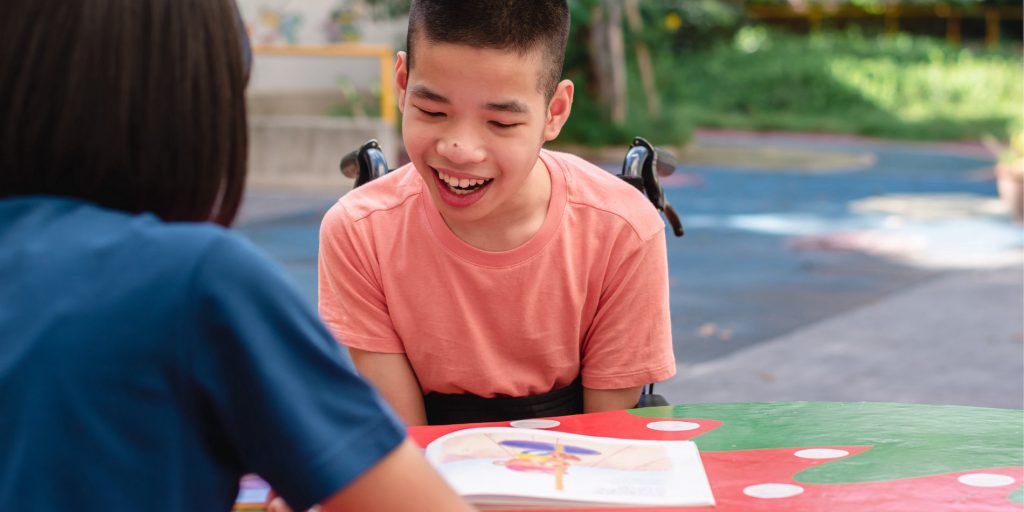 A young boy in a wheelchair looks at a book on a table in front of him