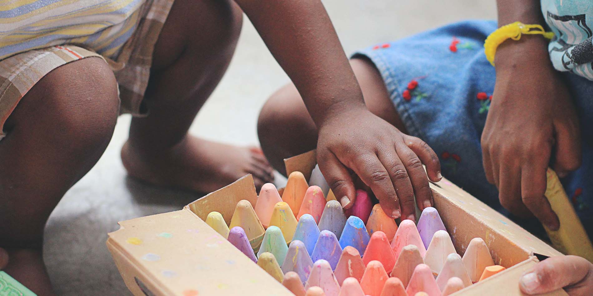 The hands of young children grab large chalk crayons that are organized in a cardboard box